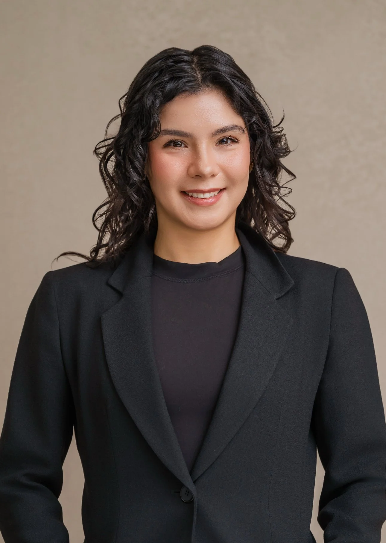 A young woman with curly dark hair styled in loose waves, wearing a black blazer over a black top, smiling against a neutral beige background.