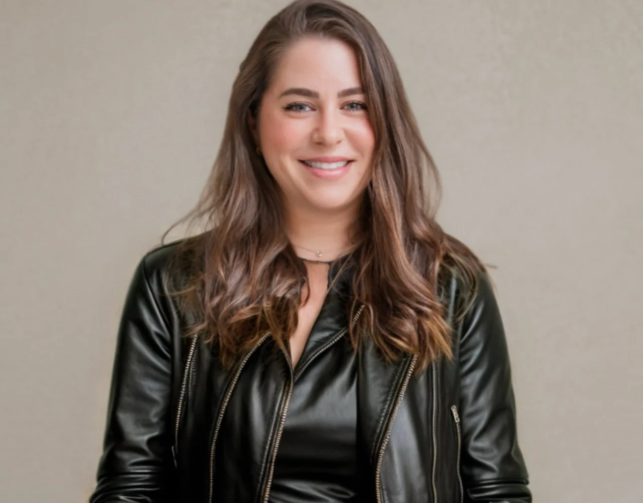 A young woman with long, wavy brown hair, smiling, wearing a black leather jacket and a black top, standing against a plain beige background.