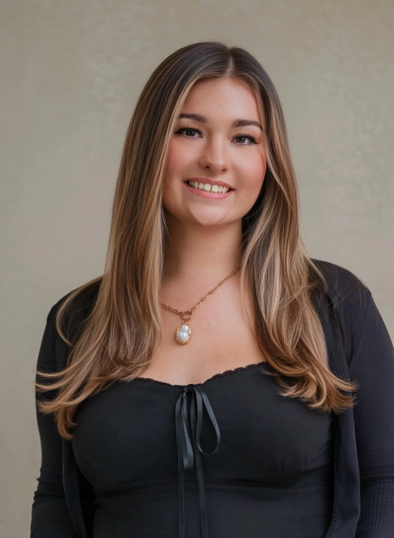 Portrait of a young woman with long, wavy brown hair and a warm smile, wearing a black top with a ribbon tie, a gold necklace with a large pearl pendant, and a black jacket, standing against a plain beige background.