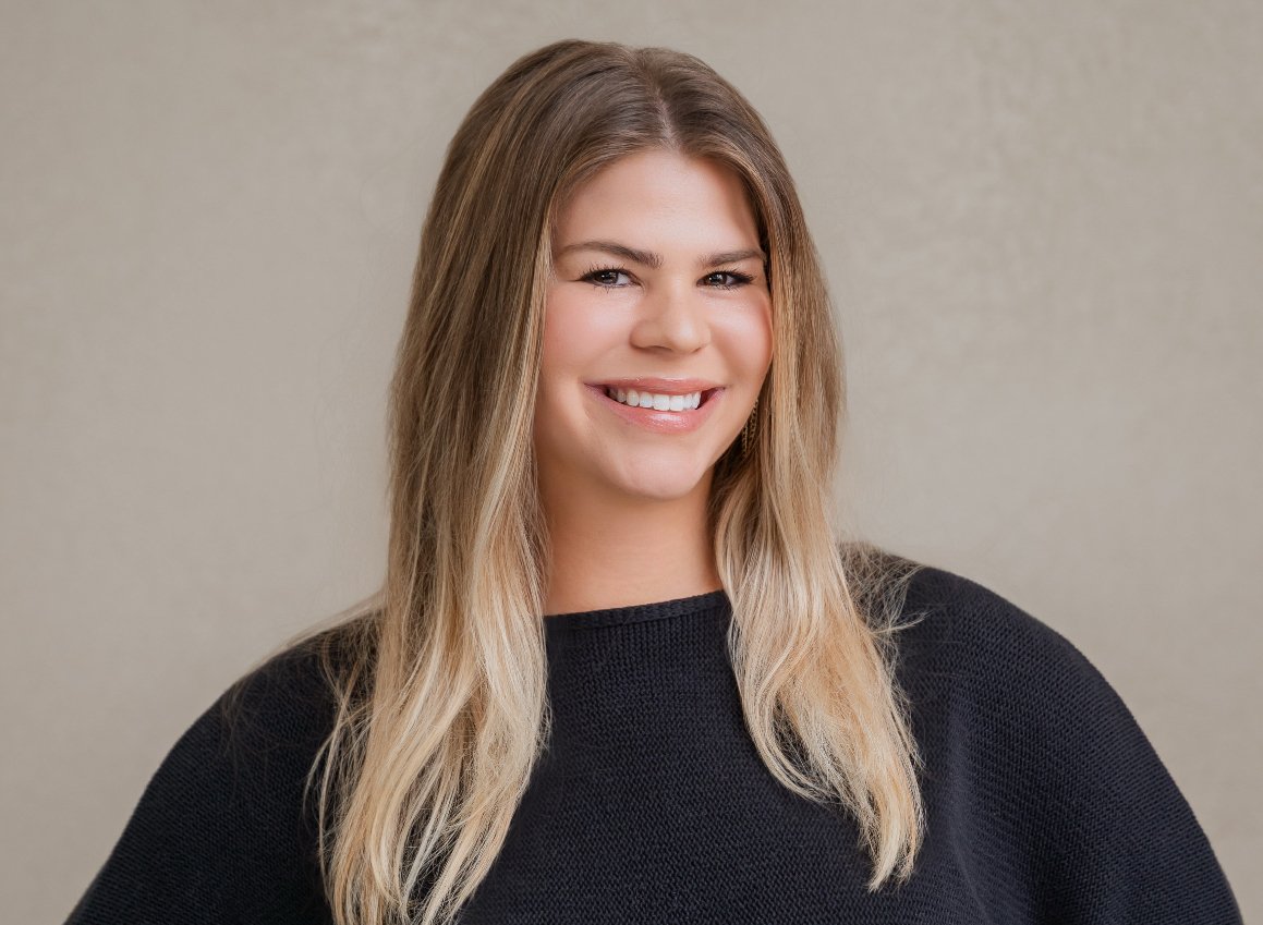 A smiling woman with long blonde hair wearing a black top, standing against a plain beige background.