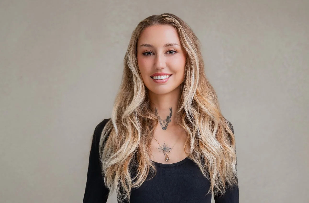 A young woman with long blonde wavy hair, wearing a black top, smiles at the camera, standing against a plain light-colored background.