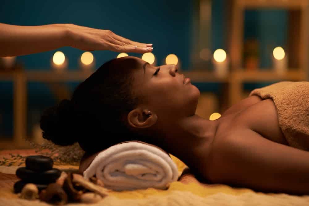 A woman receiving a massage in a spa or massage therapy setting, lying face-up on a massage table with a rolled towel under her neck, with candles and shelves in the background.