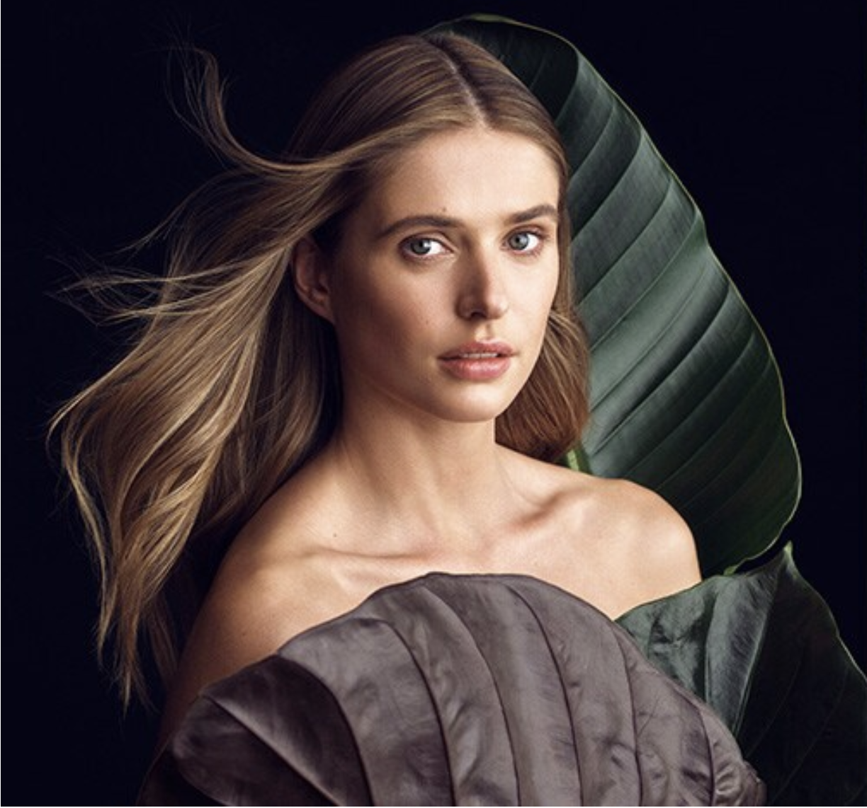 Portrait of a woman with flowing hair, looking at the camera, with large green and purple leaves behind her, against a dark background.