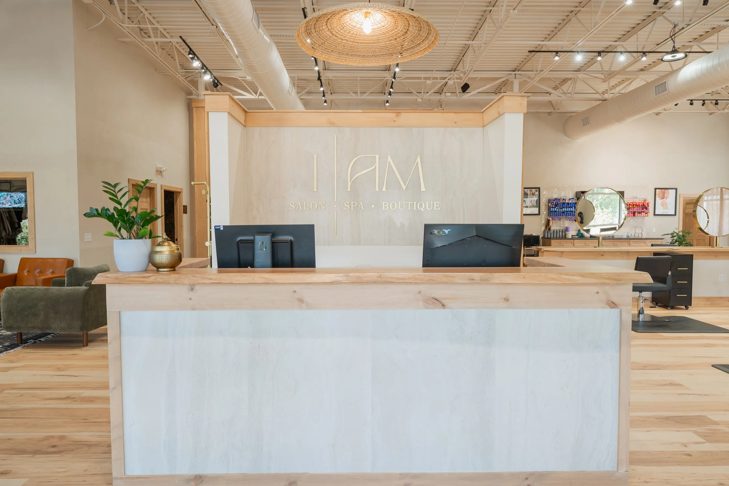 Reception area with a light wood front desk, two computer monitors, and a potted plant. To the right, there are styling stations with mirrors and supplies, and seating areas with chairs and frames on the walls. The ceiling has exposed ducts and a large woven light fixture.
