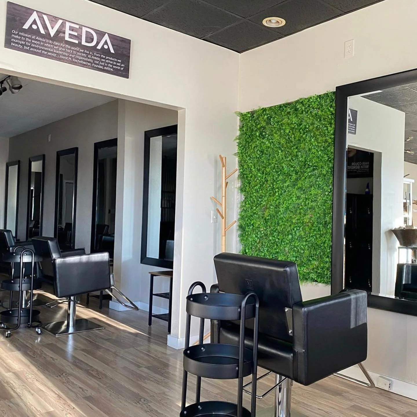 A modern hair salon with black styling chairs, large rectangular mirrors, a green decorative wall, and wooden flooring.