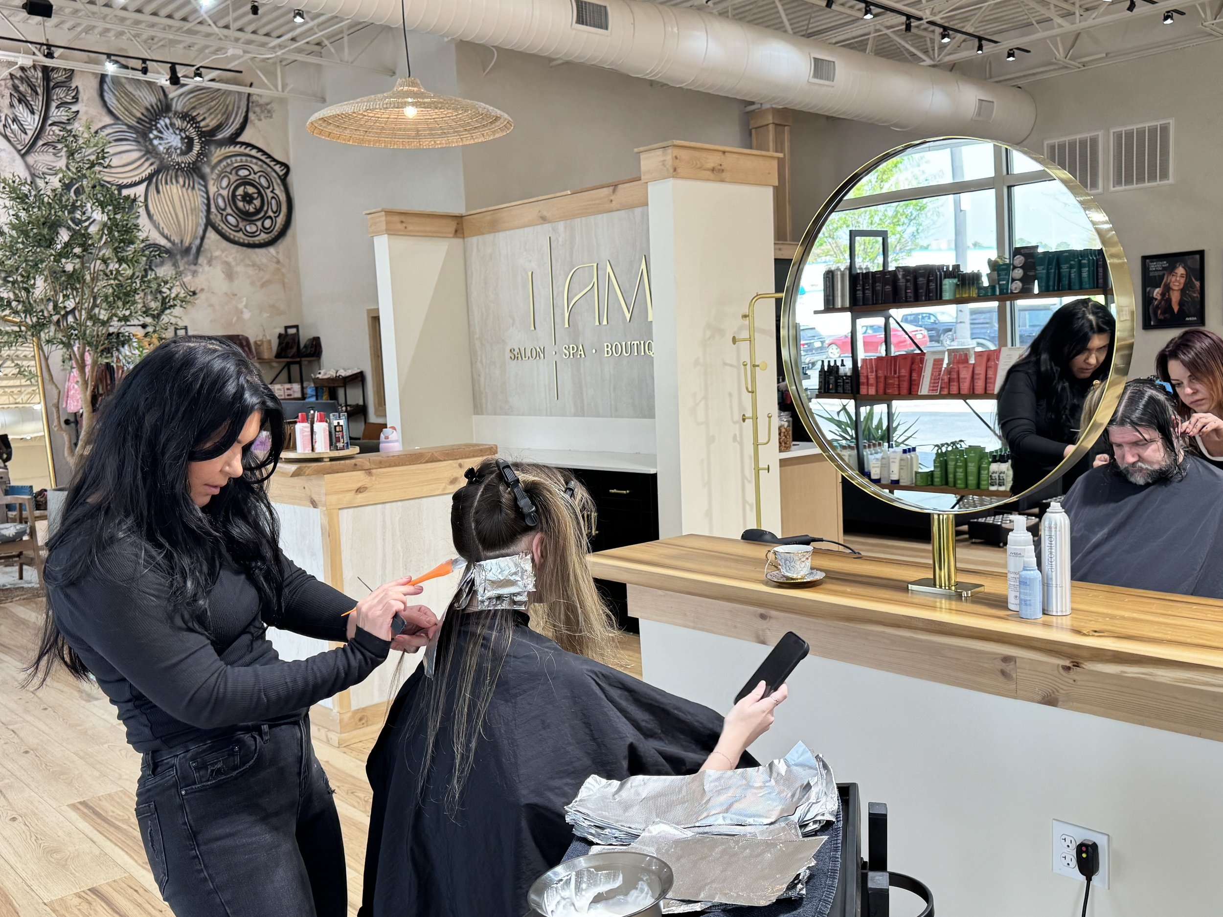 Woman getting her hair colored at a salon, sitting in front of a large round mirror, with a stylist applying hair dye and another stylist assisting. There are shelves with hair products and a large window with cars outside.