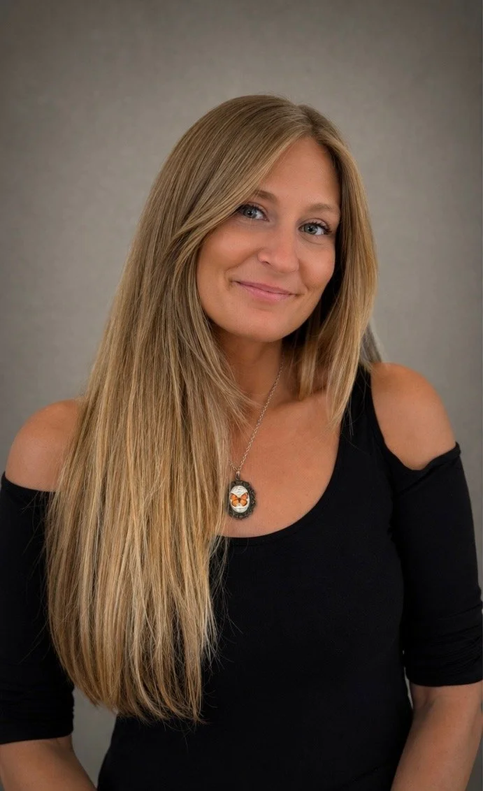 A smiling woman with long blonde hair wearing a black off-shoulder top and a necklace with a fox pendant, against a plain gray background.