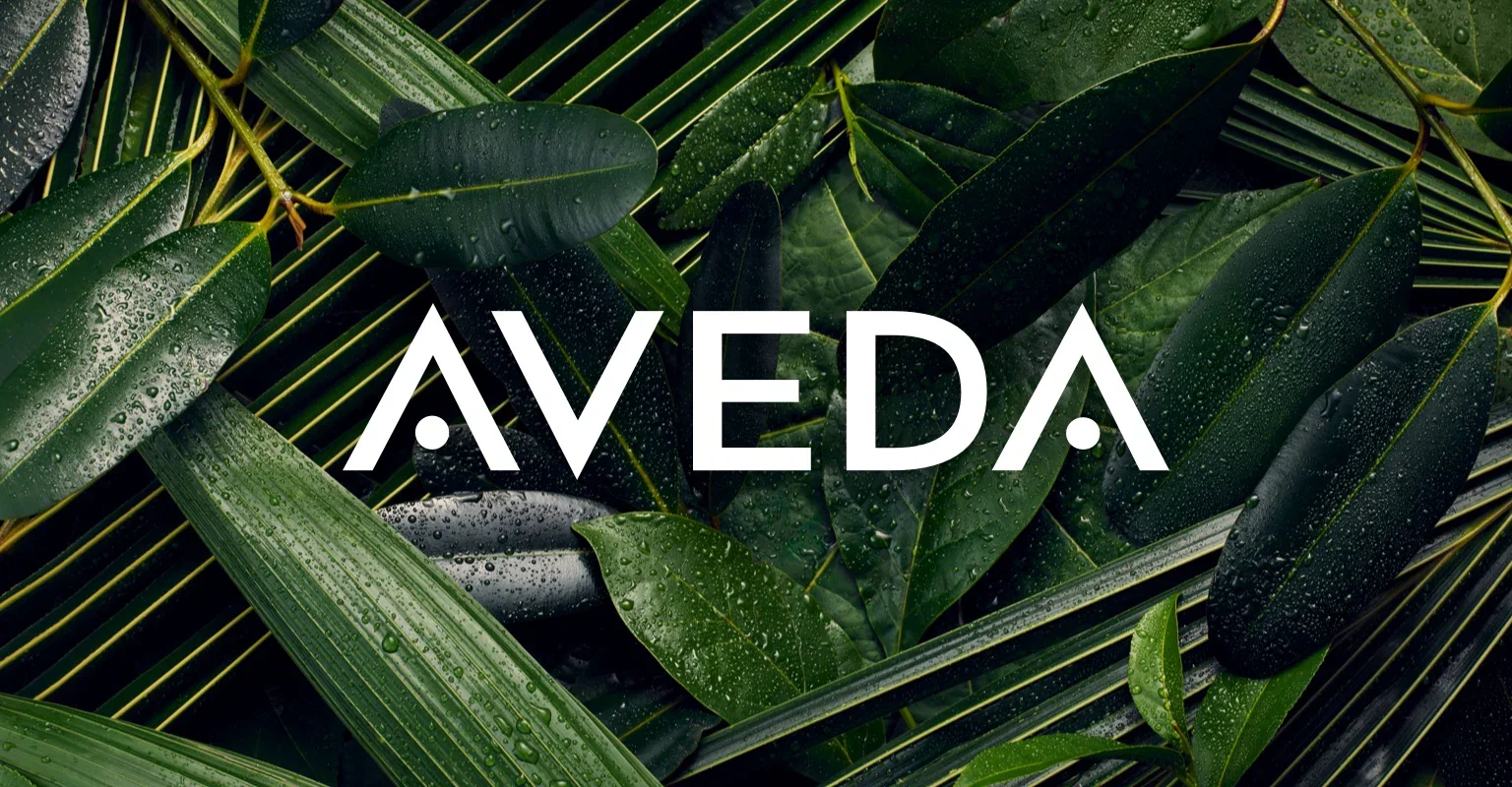 Close-up of green leaves with water droplets, with the word 'AVEDA' superimposed in white text.
