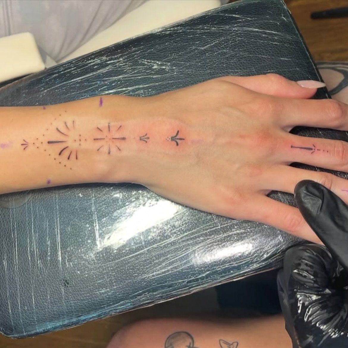 A tattoo in progress on a person's wrist and hand, with tattoo artist wearing black gloves. The tattoo includes various small arrow and geometric designs, some with dotted lines, on the top of the person’s hand and wrist.
