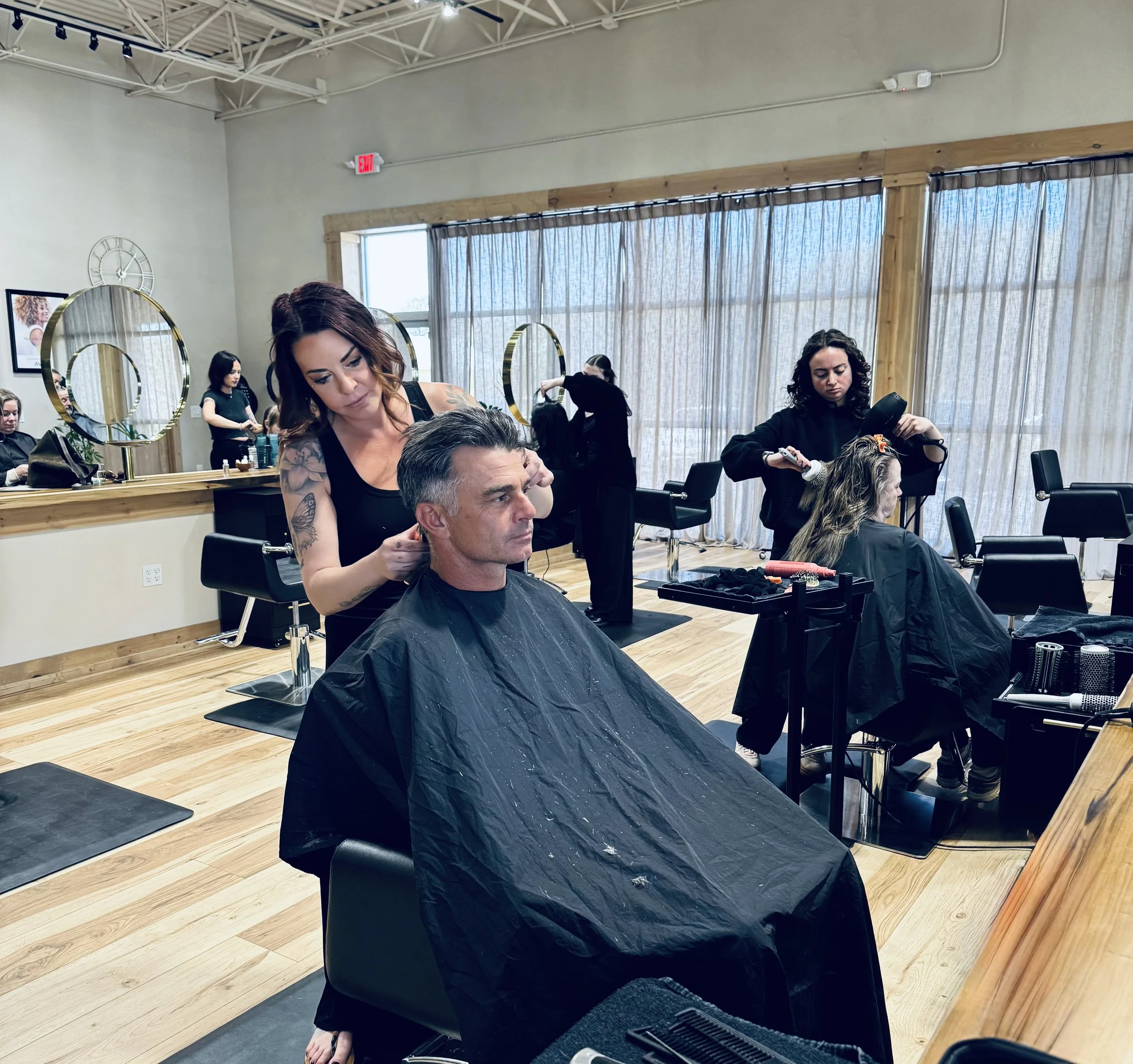 A man getting a haircut at a salon with stylists working on other clients. The salon has large windows with curtains, wooden floors, and salon chairs.