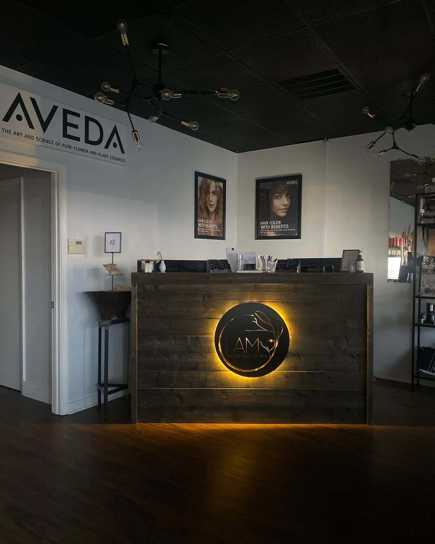 Front desk of a salon and spa named 'I AM' with a illuminated round logo on a wooden counter, with posters of hair color benefits on the wall behind, and a sign with the price $5 beside the desk.
