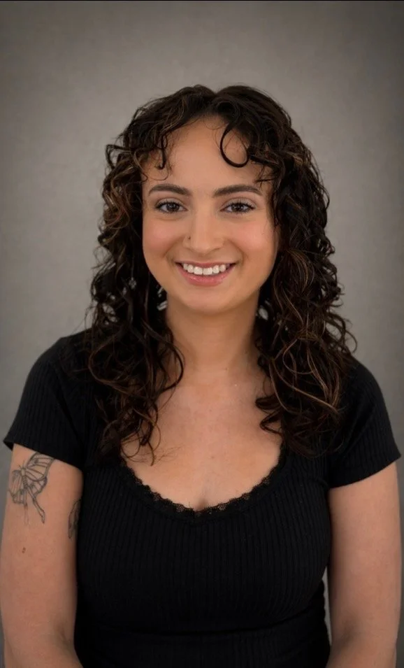 Portrait of a young woman with curly brown hair, smiling, wearing a black short-sleeve top, with a butterfly tattoo on her left arm, against a plain background.