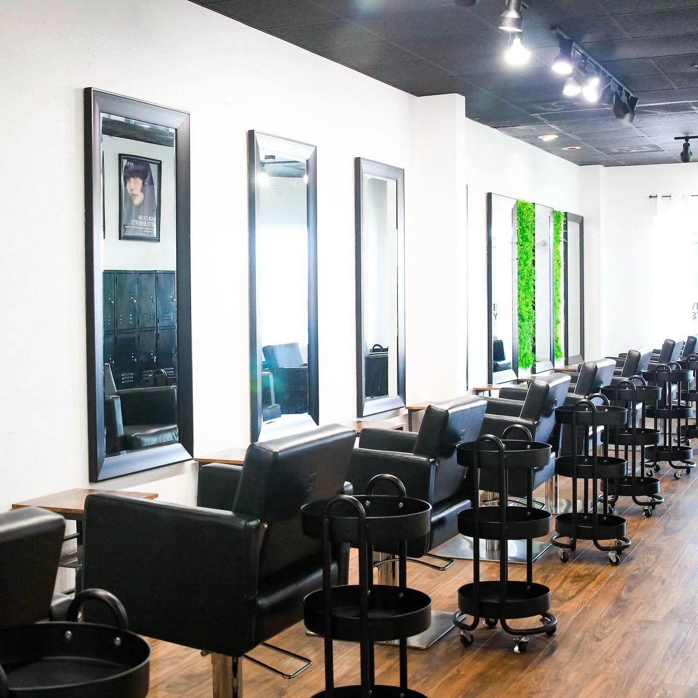 Salon with black chairs, large mirrors, lockers, and a decorative green wall, illuminated by ceiling spotlights.