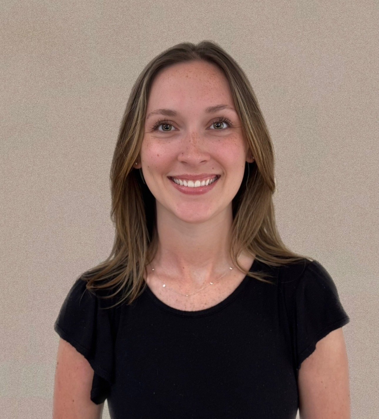 A smiling woman with shoulder-length brown hair wearing a black top and a delicate gold necklace, standing in front of a beige plain background.