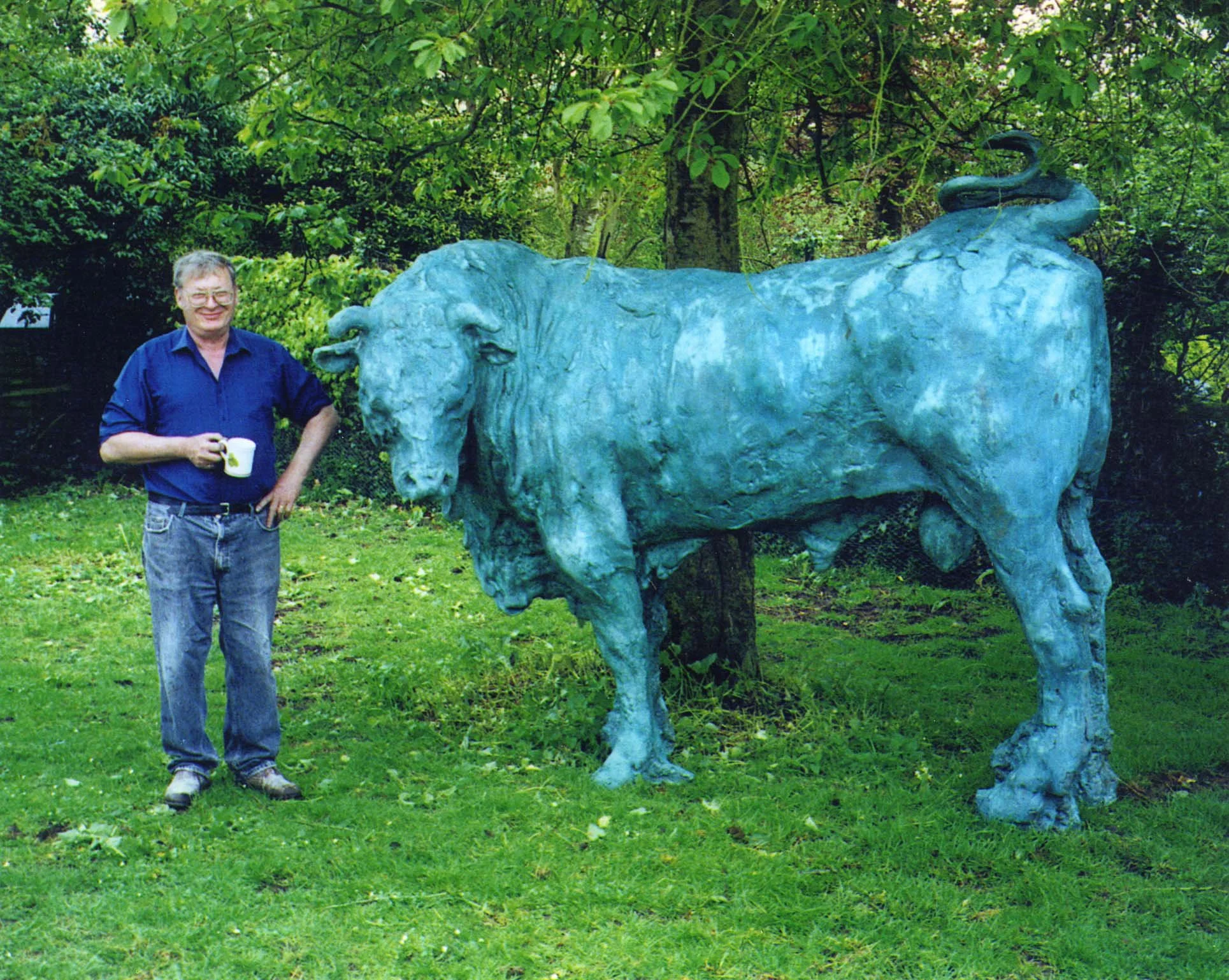 Sculptor Brian Taylor standing outdoors next to his large bronze sculpture of a buffalo in a lush green park.
