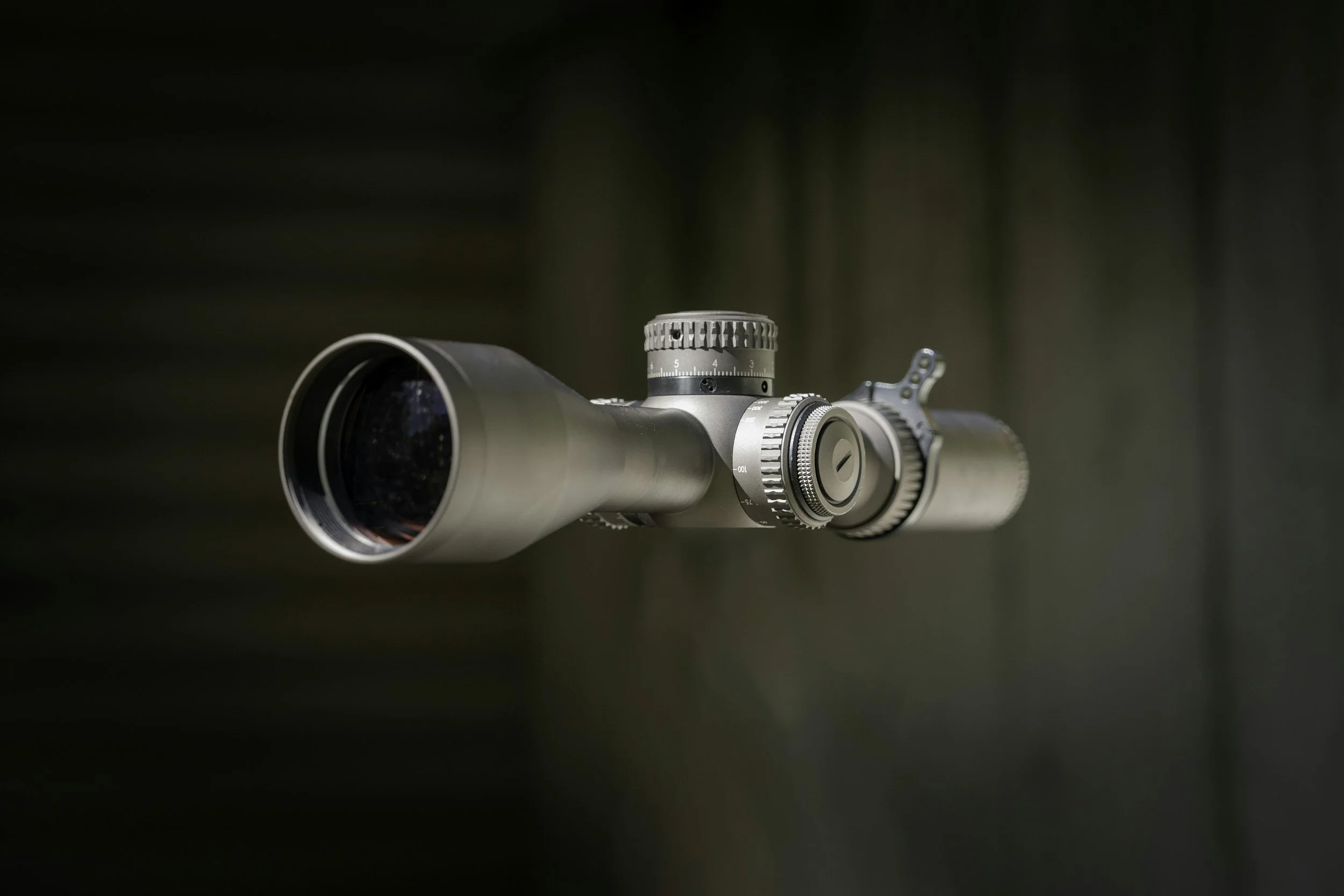 Close-up of a modern rifle scope with metallic finish and adjustment knobs, mounted on a firearm against a blurred background.