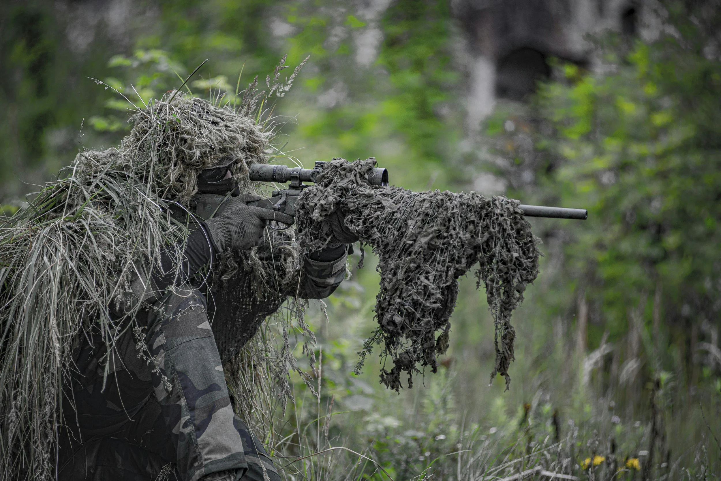 A soldier in camouflage gear and a ghillie suit aiming through a sniper scope in a forest.