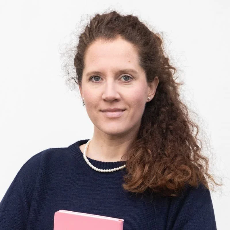 Porträt einer Frau mit lockigem braunem Haar, blauer Augen, schwarzem Pullover, Perlenkette und einem pinkfarbenen Buch vor weißem Hintergrund.