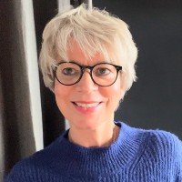 Porträt einer Frau mit kurz grauem Haar, Brille und blauem Pullover, lachend vor einem dunklen Hintergrund.