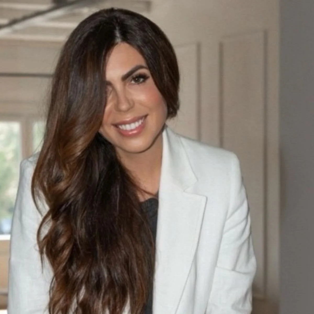 Eine Frau mit langen, braunen, gewellten Haaren trägt eine weiße Jacke und lächelt in die Kamera.