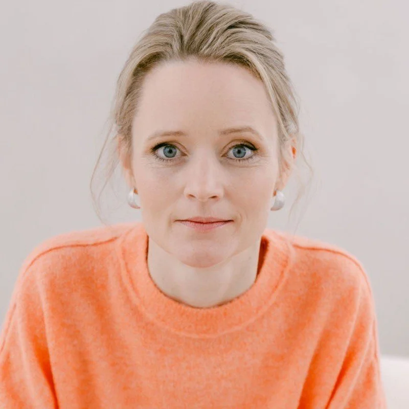 Eine Frau mit blonden Haaren, blauen Augen und einem hellen Teint trägt ein hellorangefarbenes Pullover und silberne Ohrringe, schaut in die Kamera.