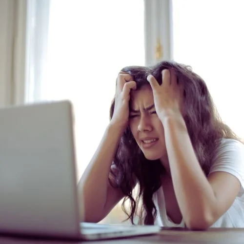 Junge Frau mit Kopfschmerzen vor einem Laptop, die die Hände im Haar hat, in einem hellen Raum mit Fenster im Hintergrund.