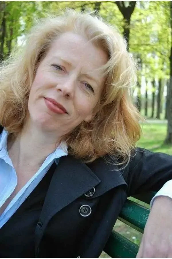 Eine Frau mit halblangem Rotblondem Haar sitzt im Park. Sie traegt eine schwarze Jacke und eine hellblaue Bluse.