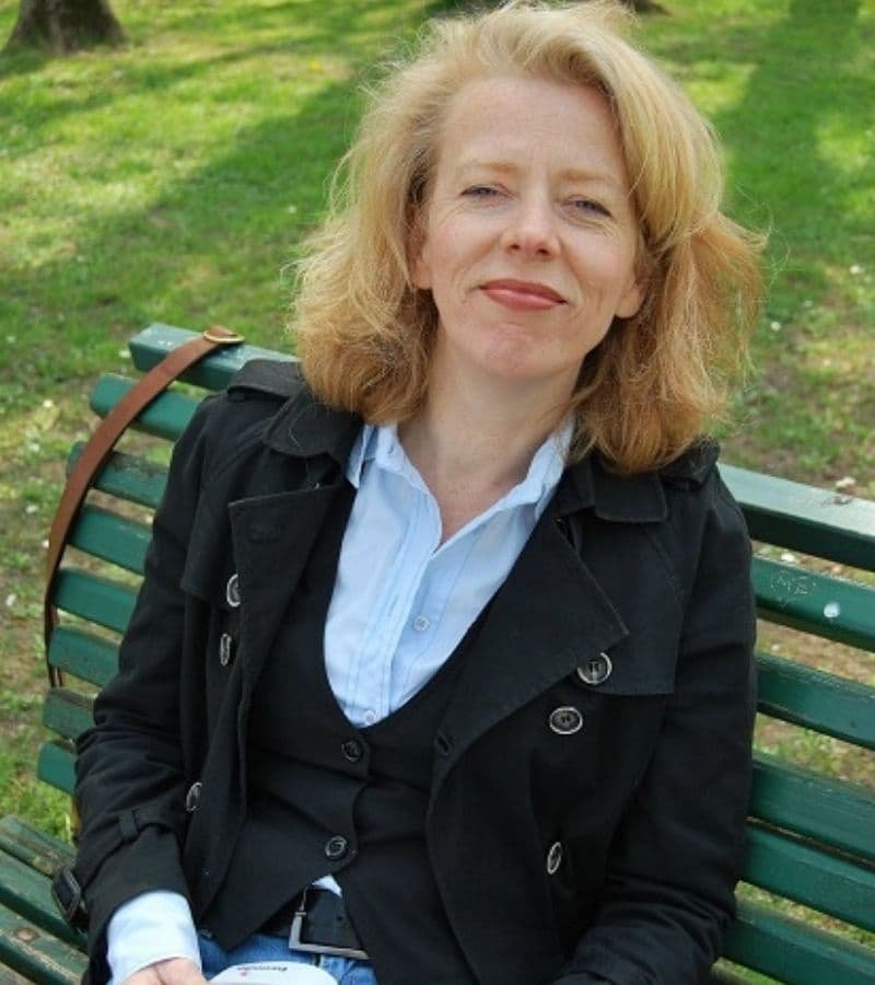 Eine Frau mit rotblonden halblangen Haaren und einer hellblauen Bluse, einer Jeans und einer schwarzen Jacke, sitzt auf einer Parkbank im Gruenen