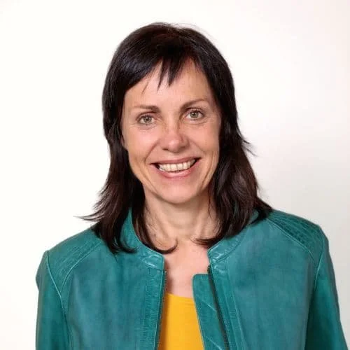 Eine Frau mit dunklen Haaren, die ein grünes Jacke trägt und lächelnd in die Kamera schaut.