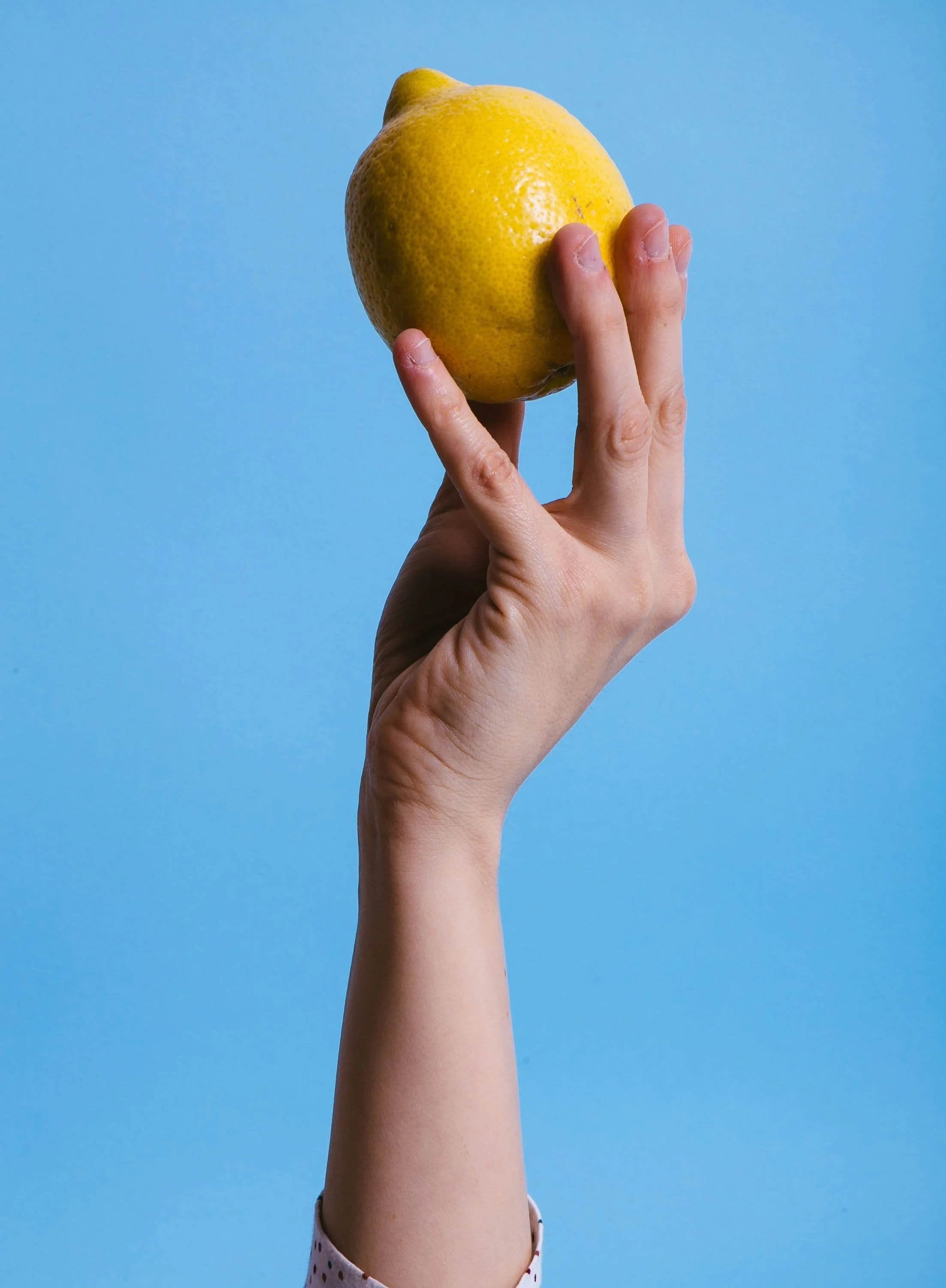 Eine Hand, die eine Zitrone vertikal hochhaelt, mit einem hellblauen Hintergrund.