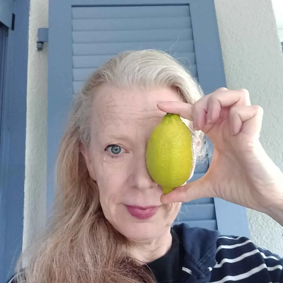 Eine Frau hält eine Zitrone vor ihr linkes Auge. Sie hat langes blondes Haar, trägt ein gestreiftes Oberteil und steht vor einer blauen Tür.