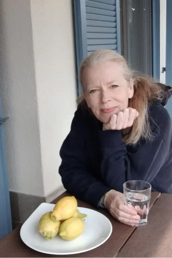 Eine Frau sitzt an einem Holztisch mit einem Glas Wasser in der Hand und einem Teller mit Zitronen vor ihr. Sie hat lange, blonde Haare und trägt einen Hoodie, blickt in die Kamera und lächelt.