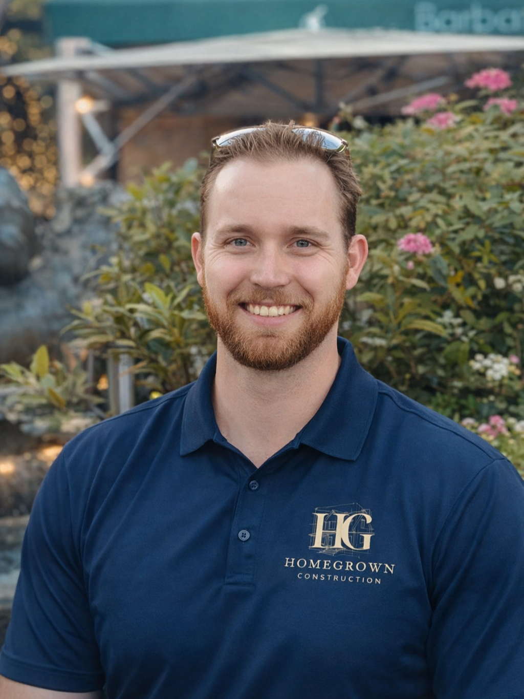 Smiling man with a beard and sunglasses on his head wearing a navy blue polo shirt that says Homegrown Construction, standing outdoors near bushes with pink and white flowers.