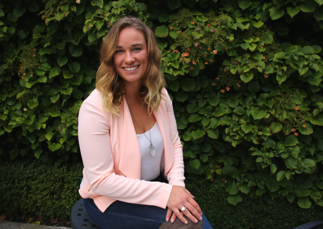 A smiling woman with shoulder-length wavy blonde hair, wearing a light pink blazer, a white top, and a silver necklace, sitting on a bench in front of a lush green hedge.