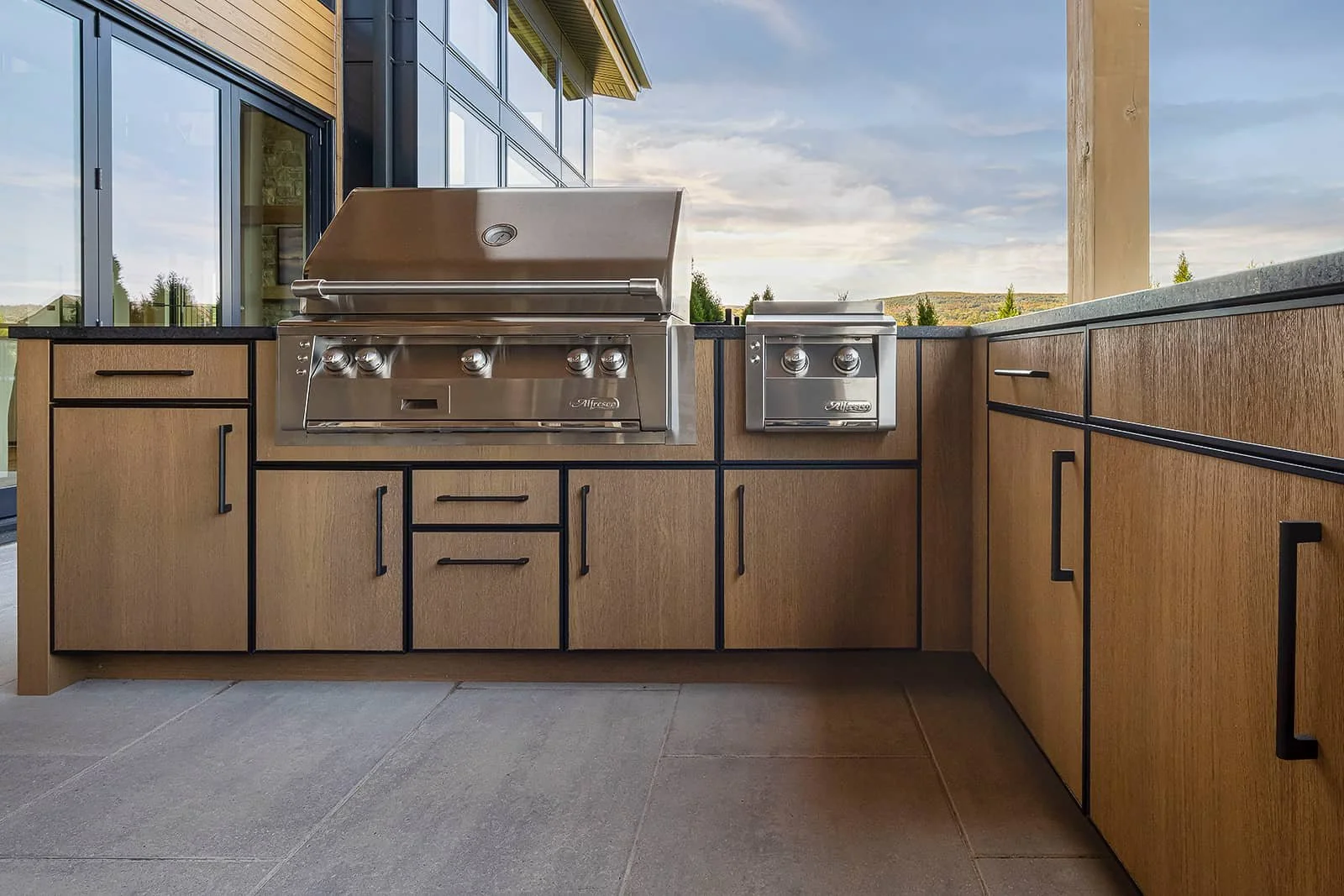 Outdoor kitchen with two stainless steel gas grills, surrounded by wooden cabinets and a black countertop, with a partly cloudy sky in the background.
