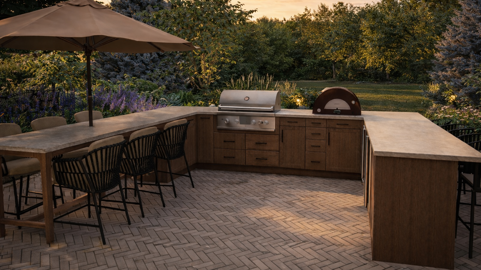 Outdoor kitchen area with a brick-patterned floor, wooden cabinets, a stainless steel grill, a pizza oven, a dining table with chairs, and a large patio umbrella, surrounded by trees and flowering plants during sunset.