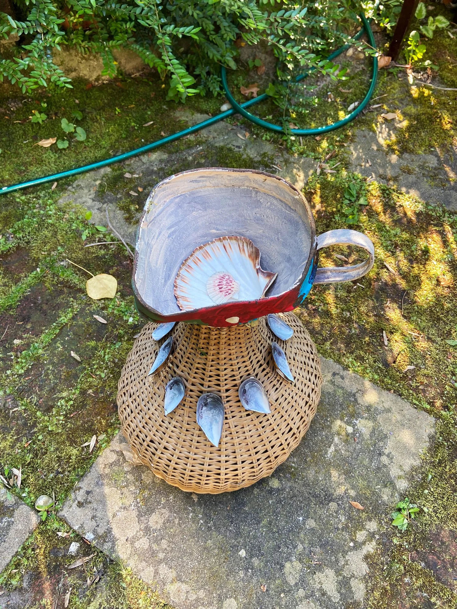 Sculptural piece made from a cup, shell, and found objects arranged on a woven base outdoors