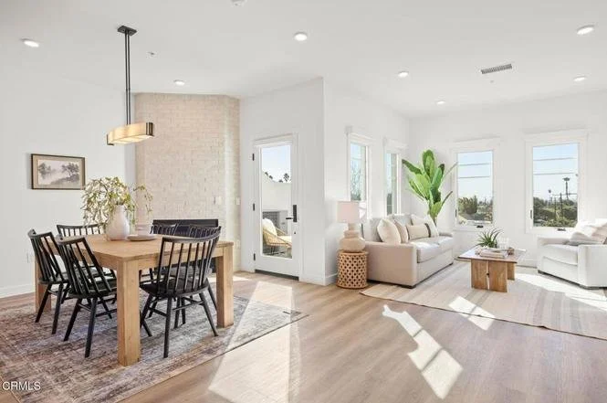Bright living room with white walls, multiple windows, and light hardwood floors. There is a cream-colored sofa, a wooden coffee table, and potted plants. Adjacent to the living room, a dining area features a wooden table with six black chairs and a hanging pendant light.