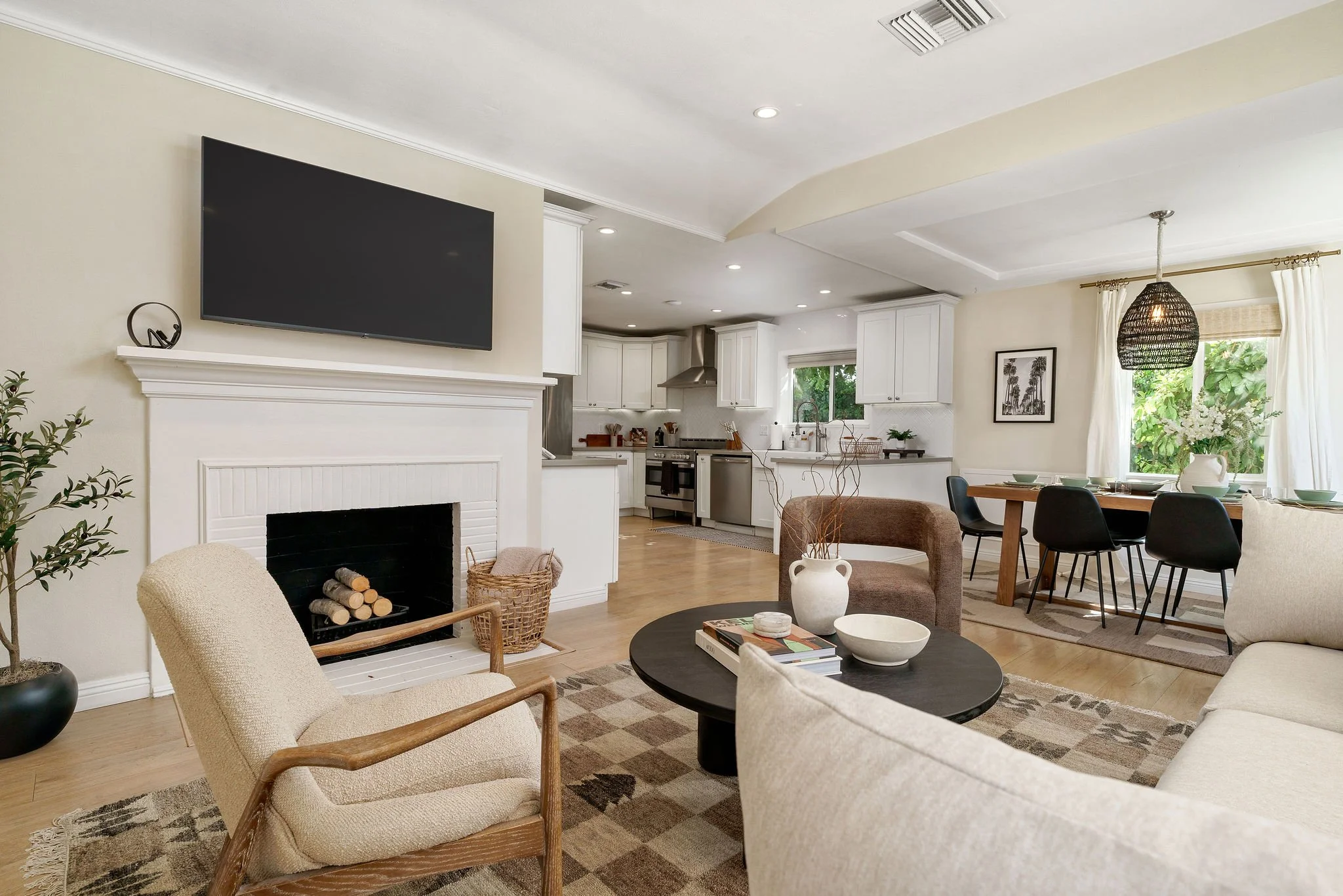 A living room with a fireplace, beige armchair, a black round coffee table with vases and books, a beige sofa, and a view into a kitchen and dining area with white cabinets, black chairs, and large windows with white curtains.