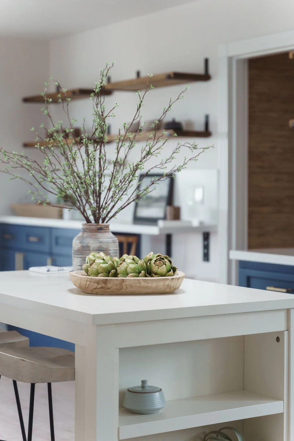A kitchen island with a white surface, decorated with a wooden bowl of artichokes and a vase of budding branches. In the background, there are blue cabinets and wall-mounted wooden shelves.