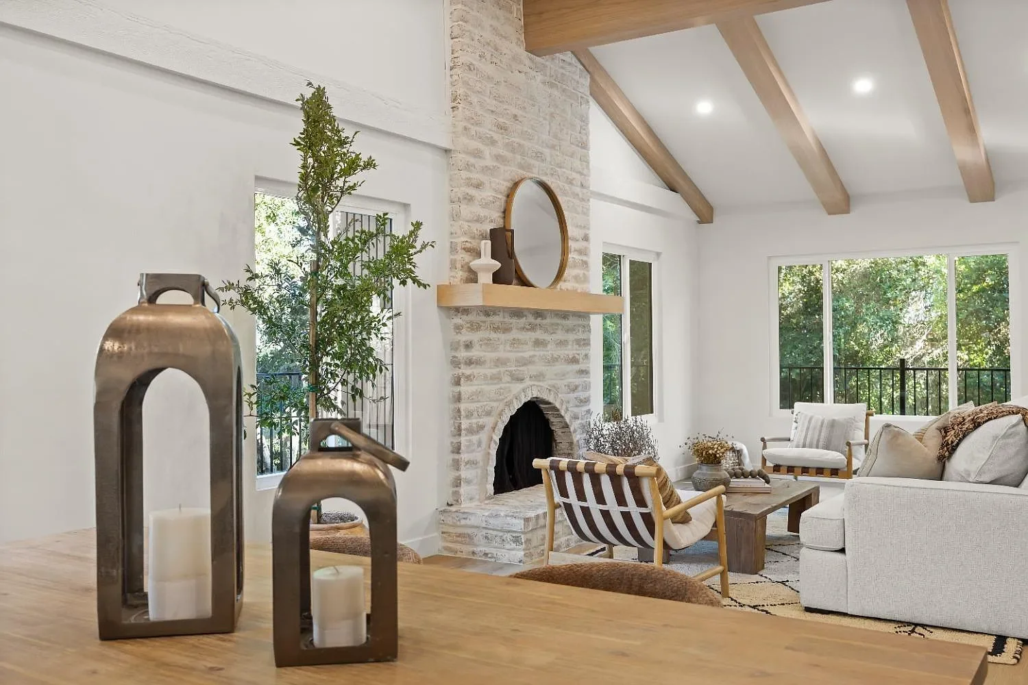 Modern living room with white walls, large windows, a light-colored sofa, armchair, fireplace with brick surround, wooden beams on the ceiling, and decorative candles on a wooden table.