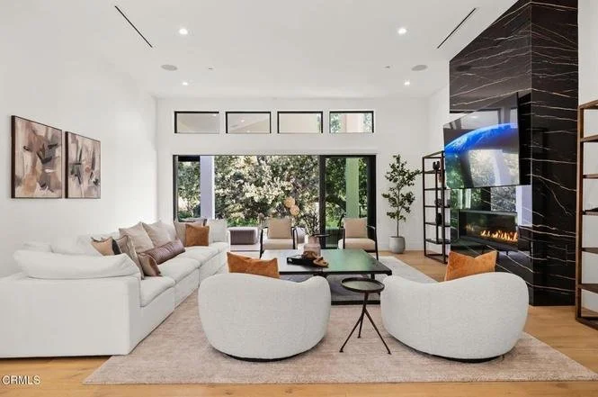 Modern living room with white sofa, two curved white chairs, a black coffee table, and a large flat-screen TV above a fireplace with black marble surround.