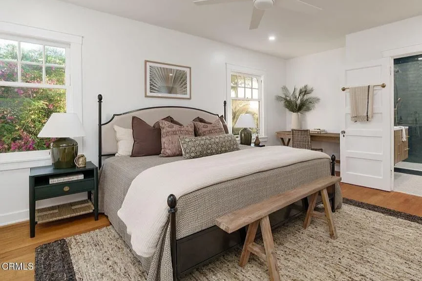 A cozy bedroom with a large bed adorned with multiple pillows, bedside lamps, windows allowing natural light, a wooden bench at the foot of the bed, a desk with a chair, and a sliding door leading to a bathroom.