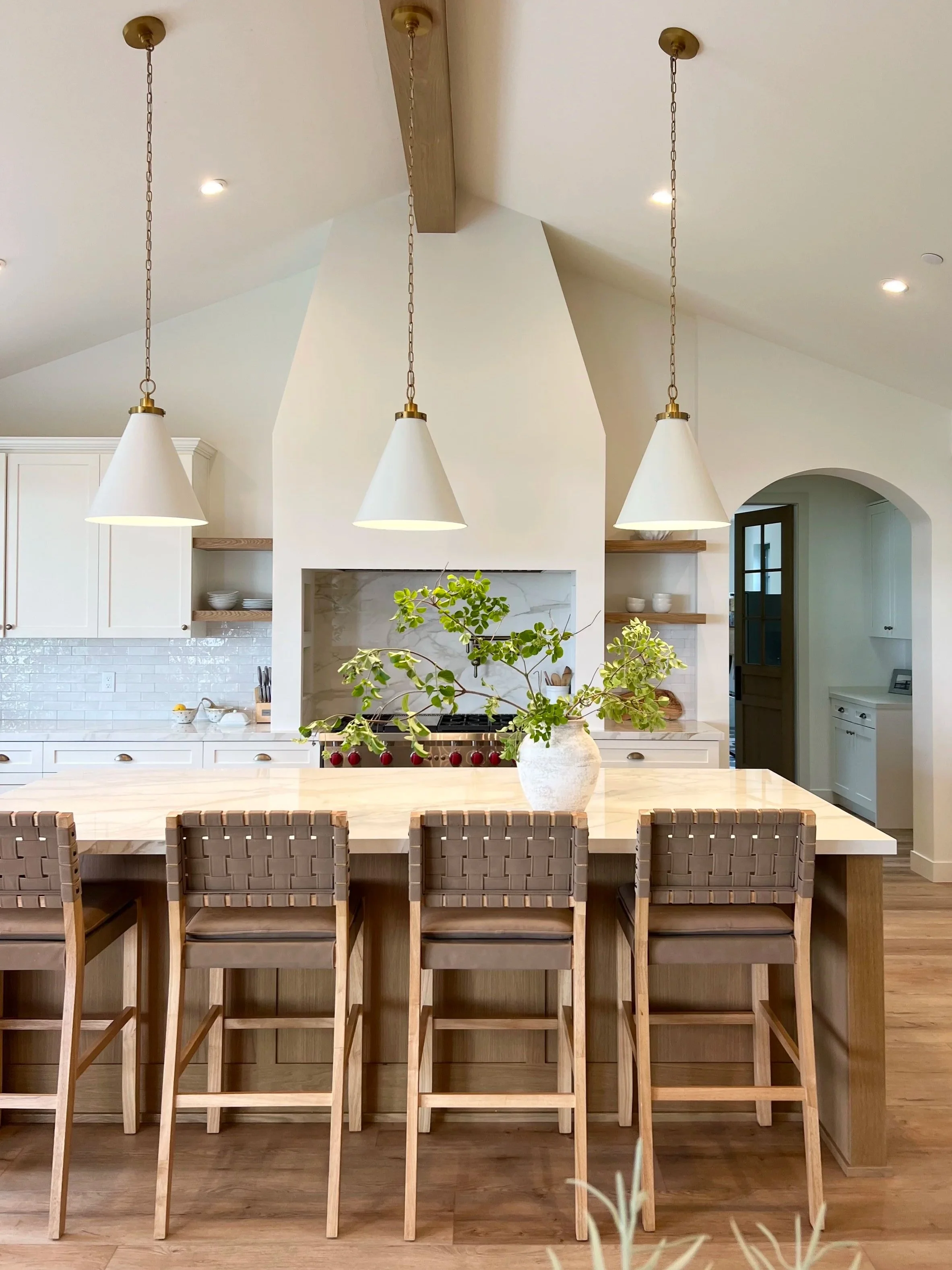Modern kitchen island with four bar stools, white vases with green foliage, pendant lights, and a stove in the background.
