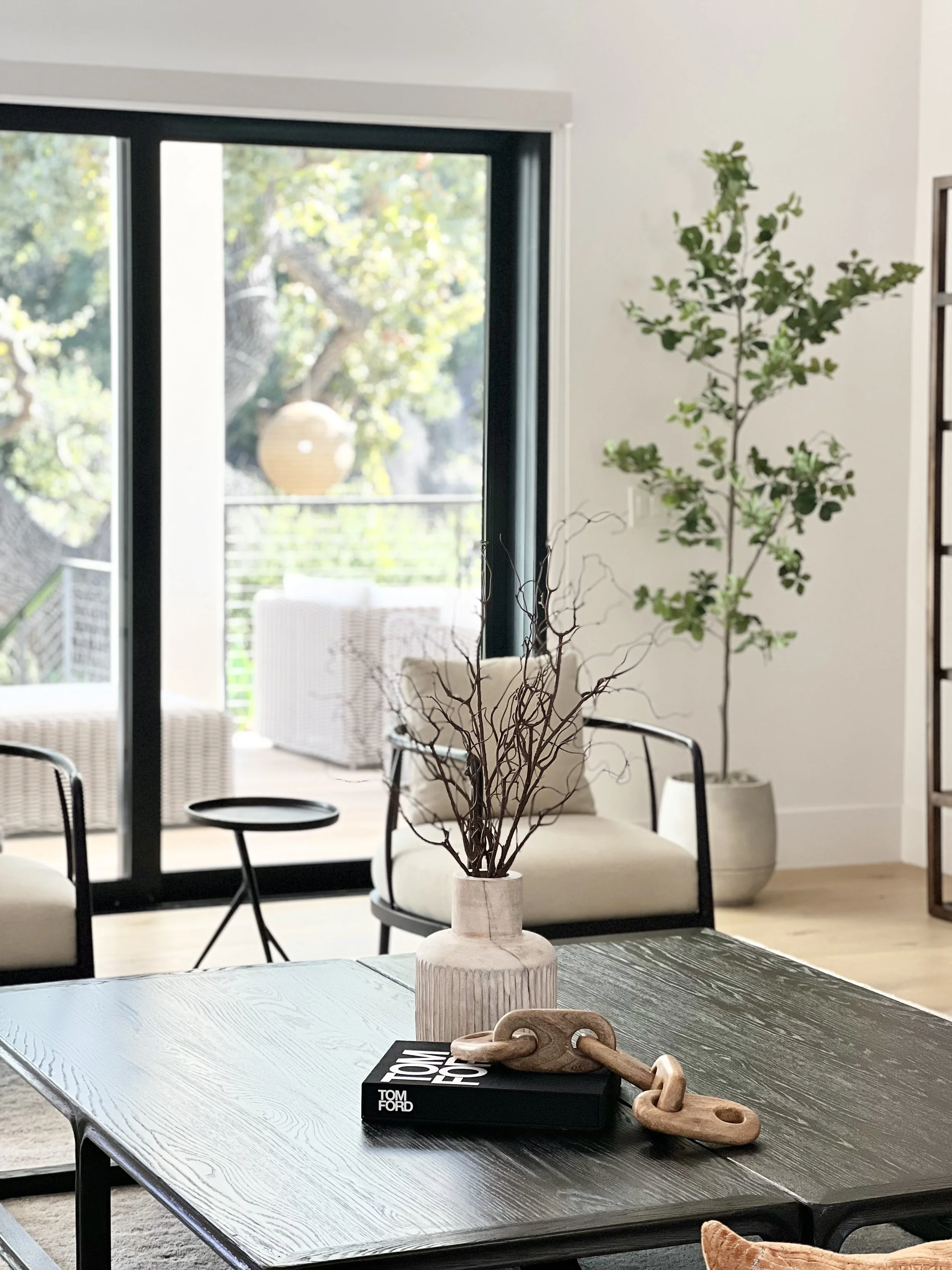 Living room with sliding glass door leading to outdoor balcony, potted plants, armchair with cushion, and black coffee table with a vase and decorative chain.