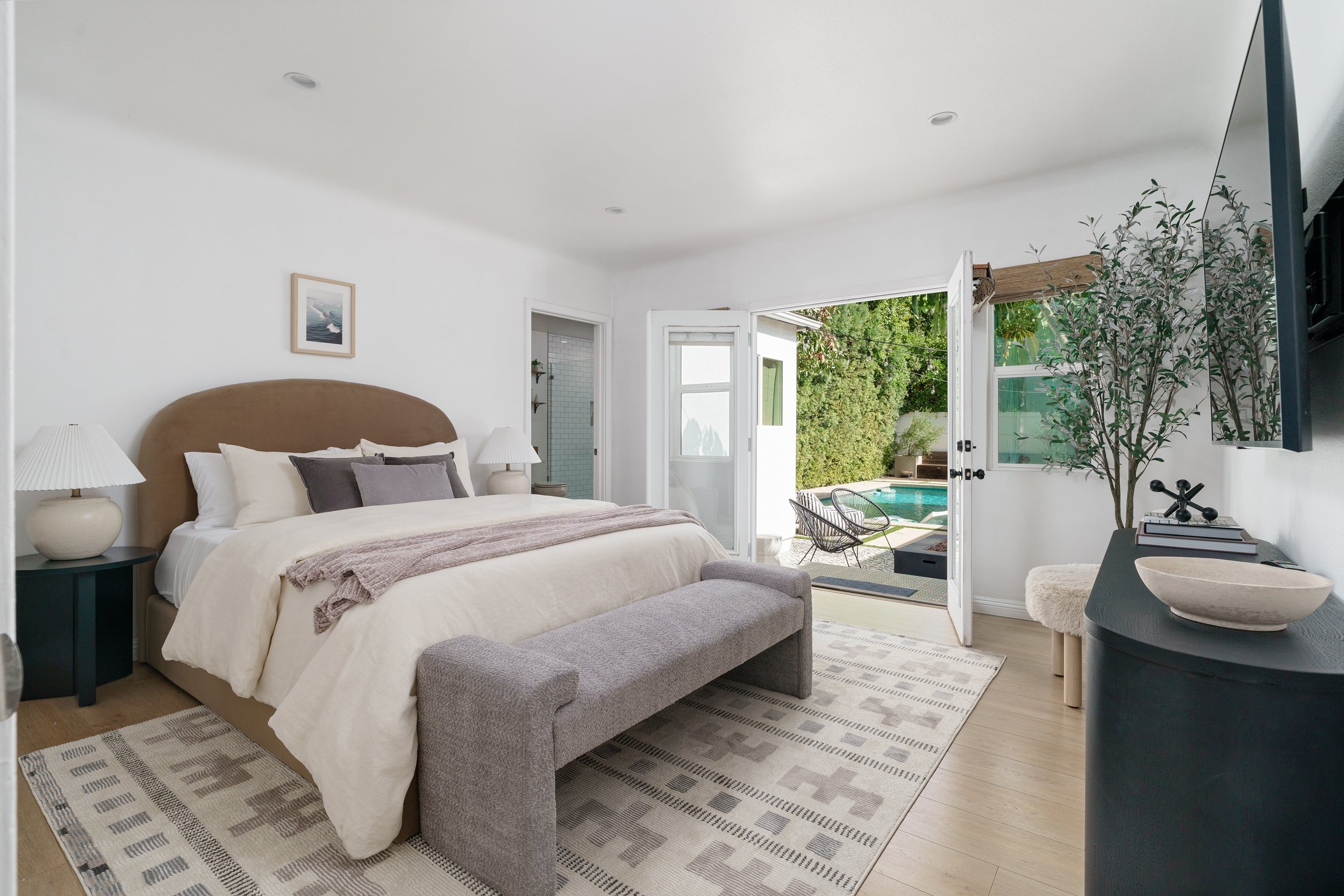 A cozy bedroom with a large bed, nightstands with lamps, an open door leading to a backyard with a pool, outdoor chairs, and lush greenery.
