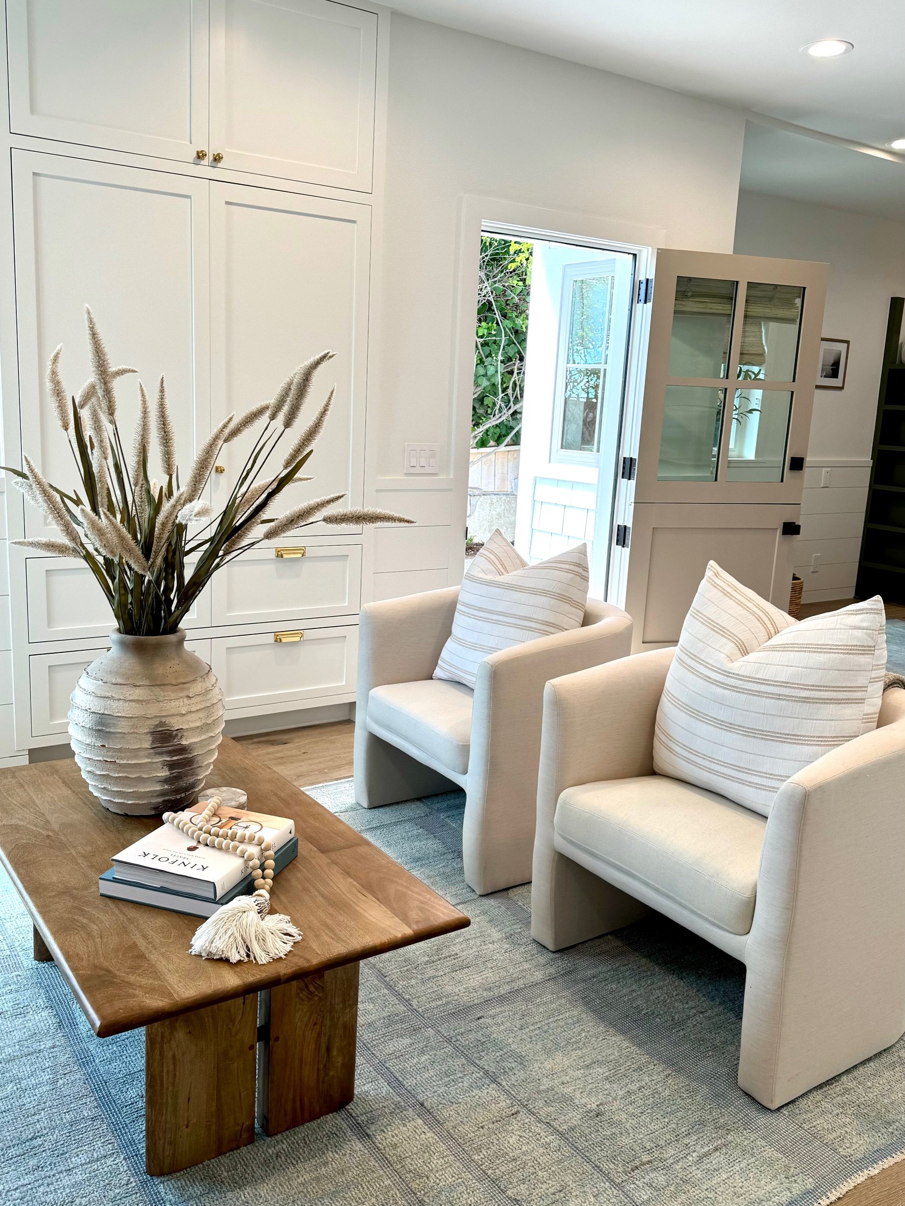 A cozy living room with two white armchairs, a wooden coffee table, a large vase with dried grasses, and an open door leading outside, with built-in white cabinets and a gray rug.