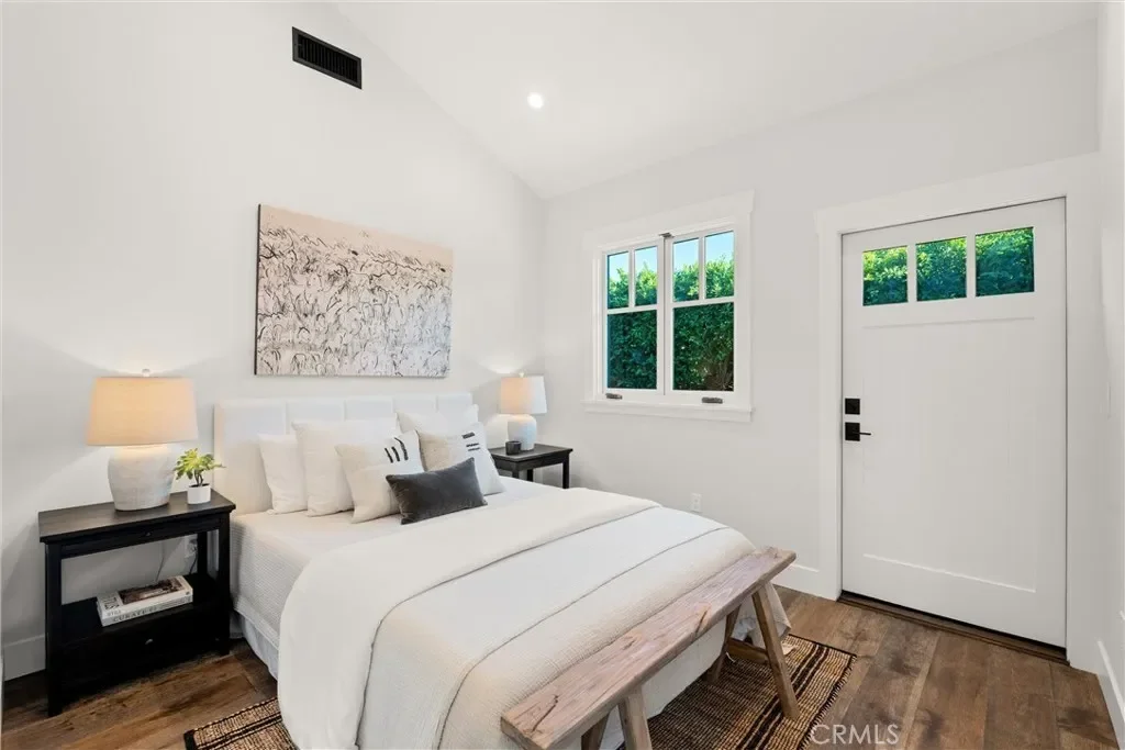 A minimalist bedroom with a white bed, dark nightstands, matching lamps, a bench at the foot of the bed, and a window with green foliage outside.