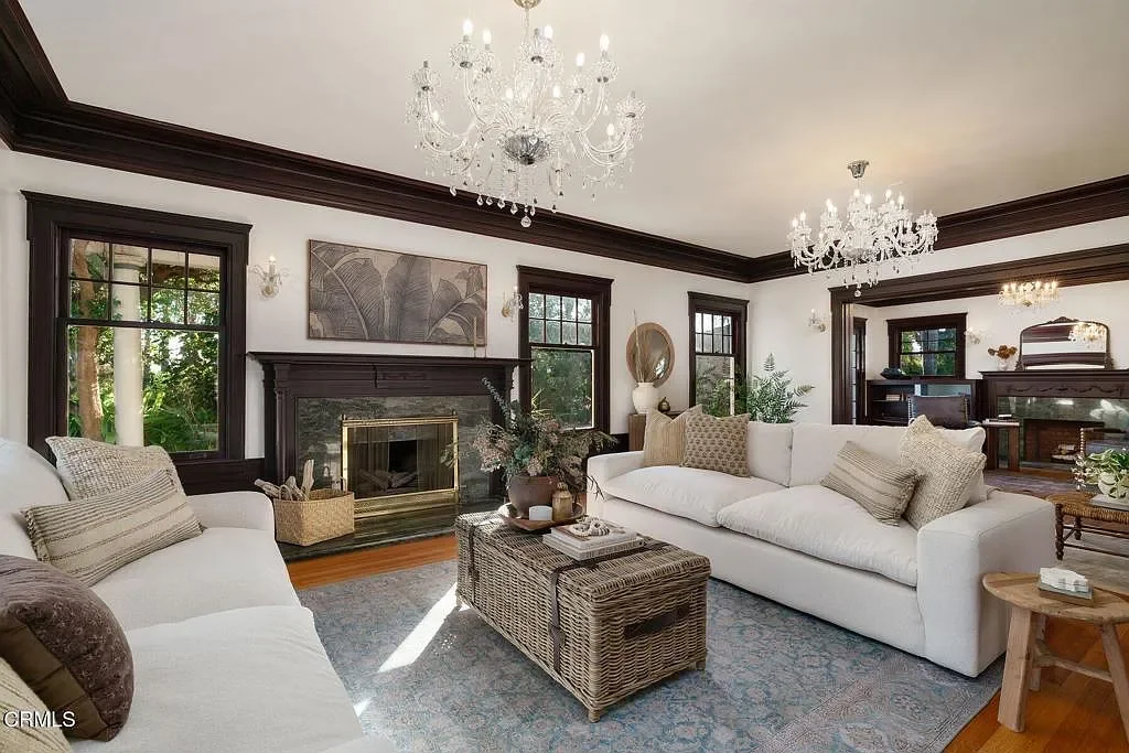 Living room with white sofas, dark wood trim, a fireplace, large windows, chandeliers, and decorative plants.