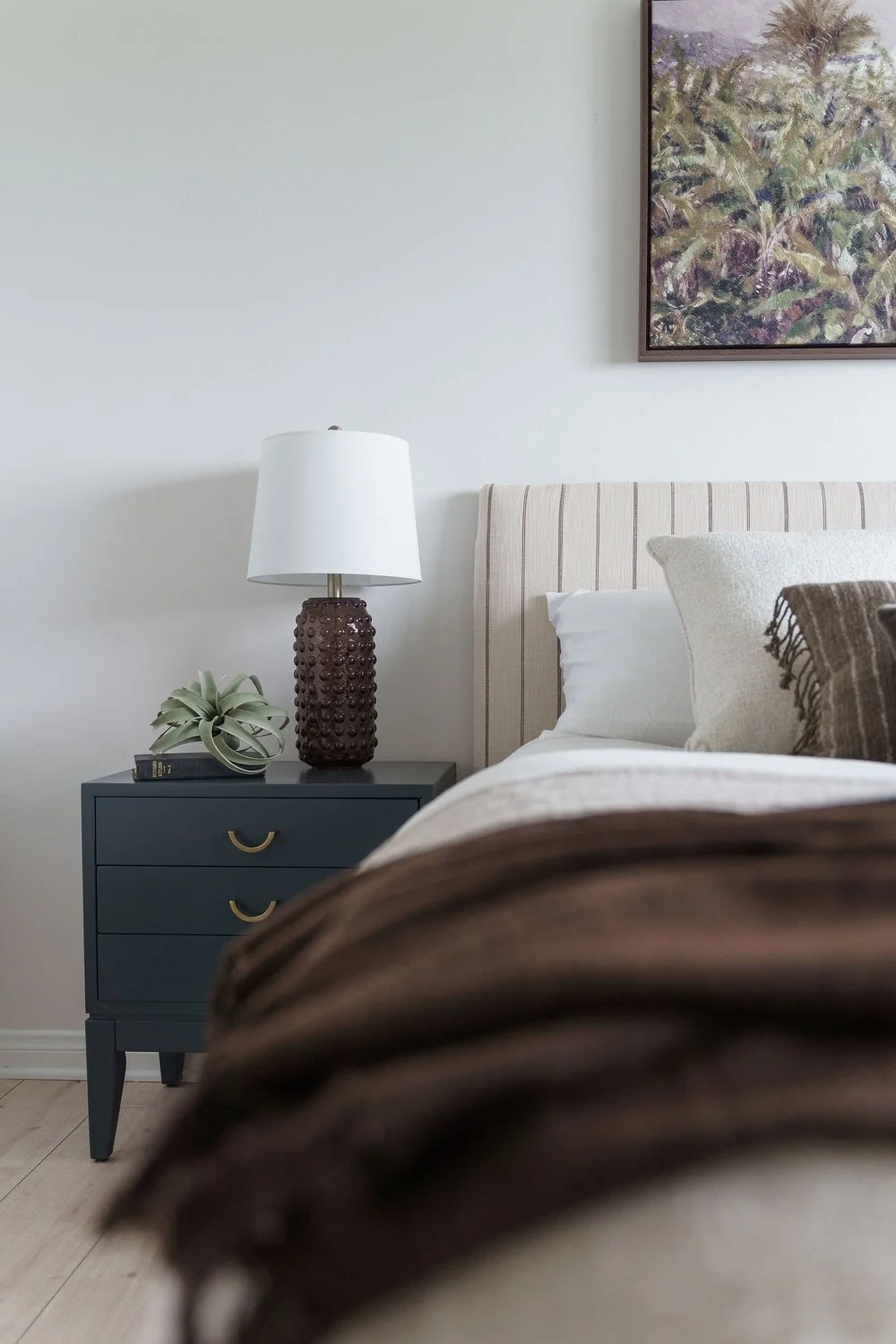 Part of a bedroom with a navy blue nightstand, a decorative lamp with a textured brown base and white shade, a small plant, a book, a beige upholstered headboard, white pillow, decorative pillow, and a framed landscape painting on a white wall.